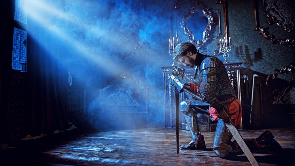 Medieval knight in armor makes an oath of allegiance by kneeling and bowing his head on the sword in the castle before the battle.