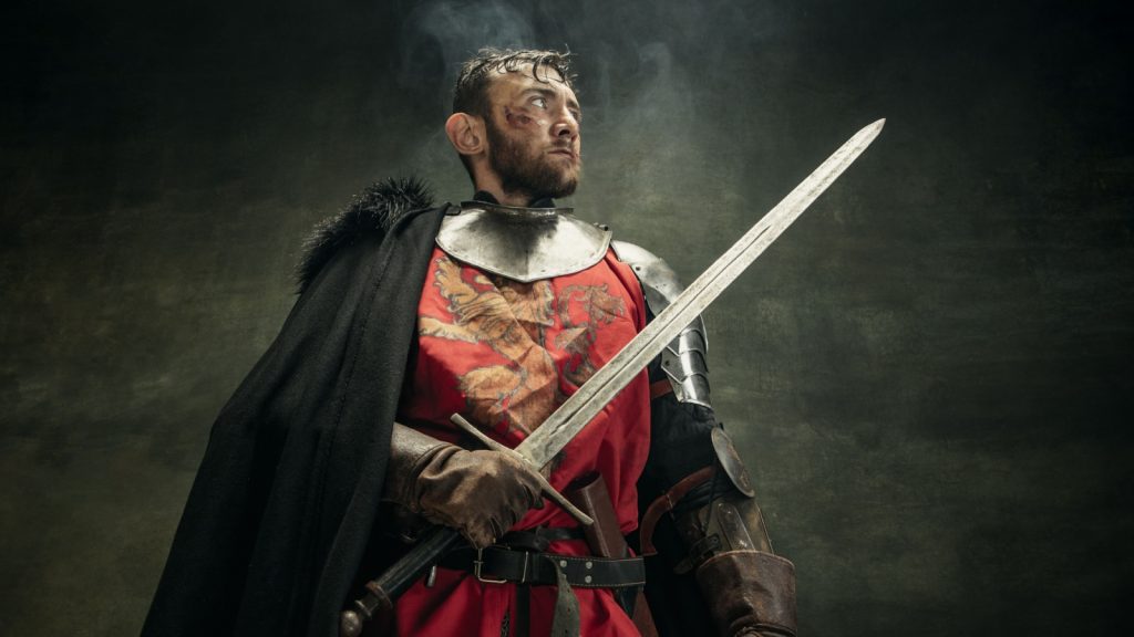 Noble warrior. Portrait of one brutal bearded man, medeival knight in cape or mantle with wounded face holding sword isolated over dark background.