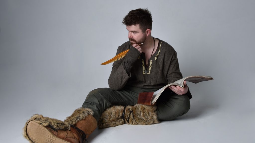 brunette man wearing historical medieval viking celtic costume, sitting pose writing in a book with quill 