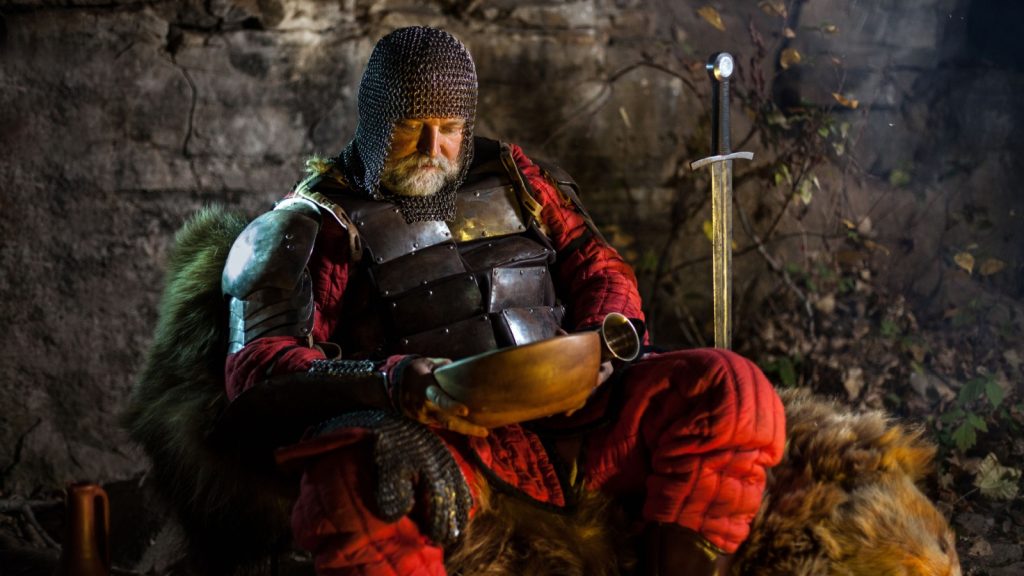 Old medieval Knight in armor with dish is sitting on furs near the camp fire and having a lunch