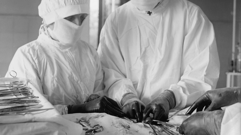 Assisting surgeon and surgical nurse with specialized instruments during an operation. Blood can be seen in surgical area. 1922 Plombage