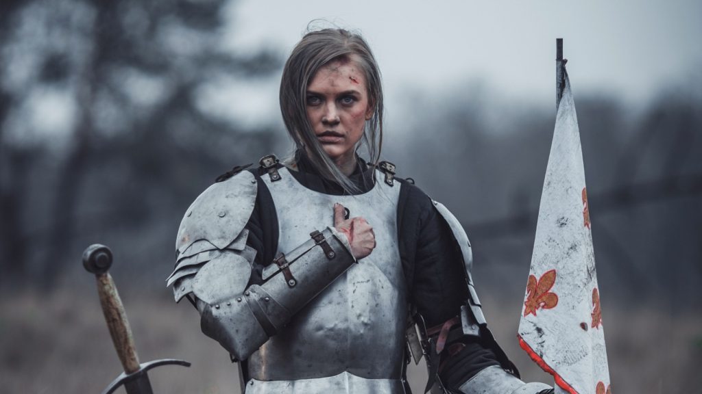Girl in image of Joan of Arc (Jeanne d'Arc) in armor kneels with flag in her hands and sword on meadow against background of dry grass.