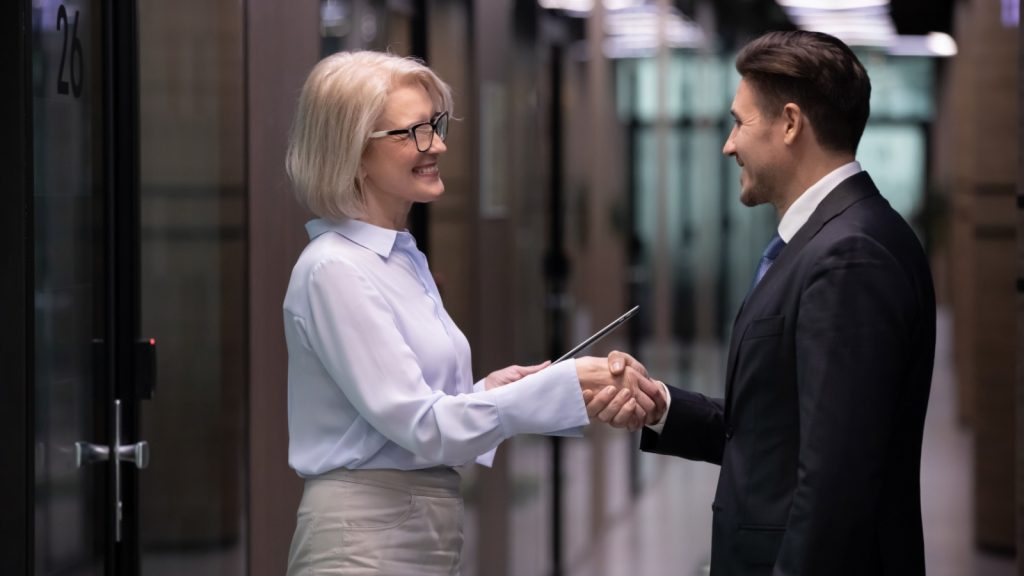 Older businesswoman and businessman shake hands standing in office. Two business partners greeting happy smiling. Senior lady boss handshaking new worker, client or colleague. Partnership handshake