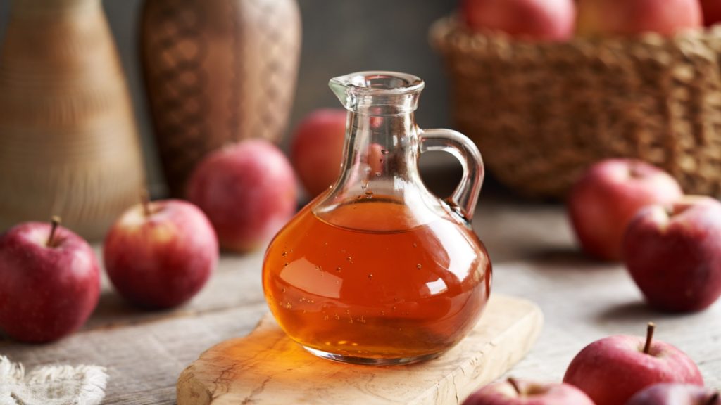 A glass jug of apple cider vinegar with fresh fruit on a table.