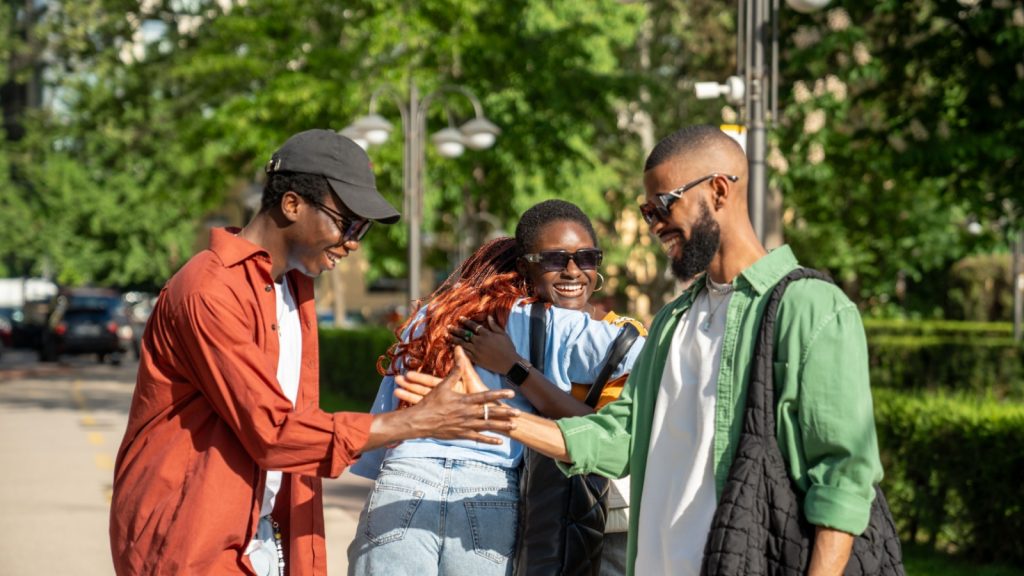 Joyous hello of african american friends on street. Happy group of black students with good mood embrace each other as meet to spend time together.Two guys shake hands, girls smile hug, see each other.