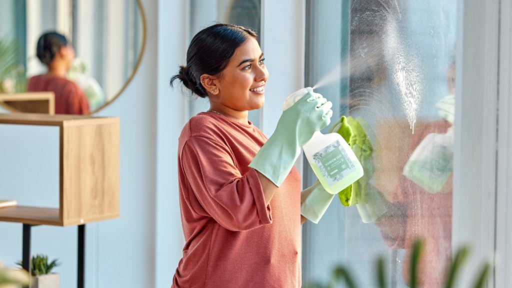 Woman, cleaning and spray product of a cleaner washing a window with a smile in home. Happy working maid use a home hygiene bottle and cloth to disinfect the house furniture with a sanitizer bottle.