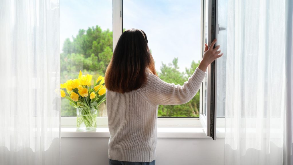 Pretty young Asian woman opening window at home on sunny day, 