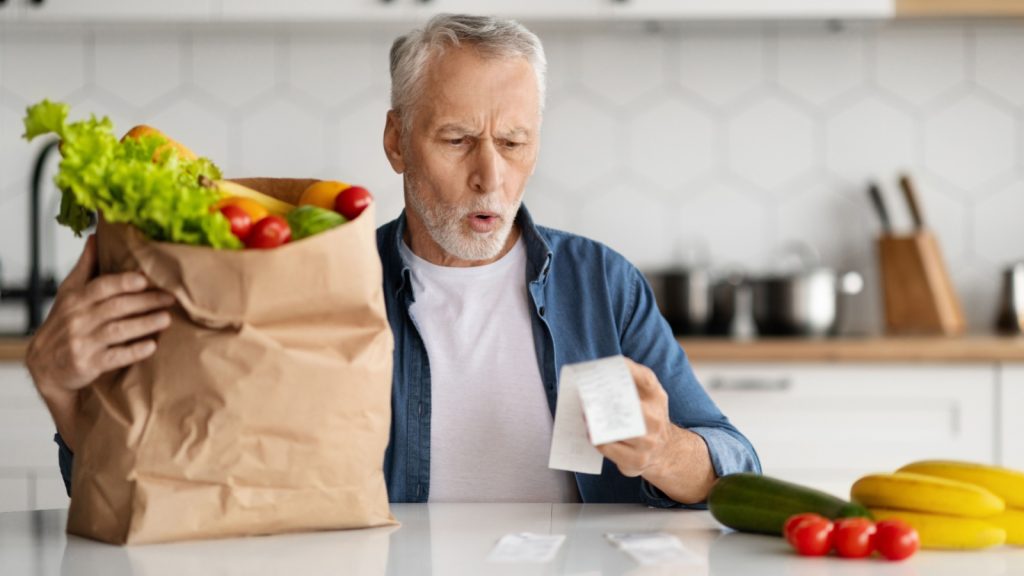 Prices For Food. Portrait Of Shocked Senior Man Checking Grocery Bills In Kitchen After Shopping In Supermarket, Frustrated Elderly Gentleman Reading Receipt With Open Mouth, 