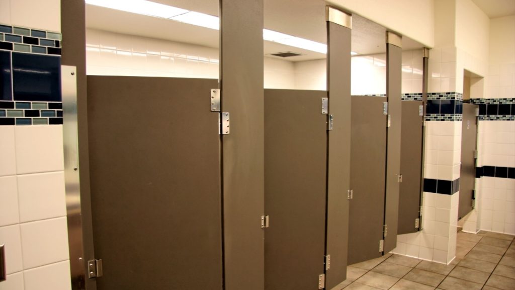 Public bathroom stalls. Four open brown metal stall doors. Fluorescent lighting in background. Beige tile floor and walls.