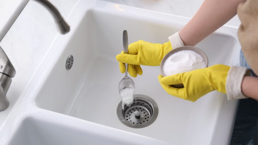 A person wearing yellow gloves pours baking soda into a sink drain using a spoon.