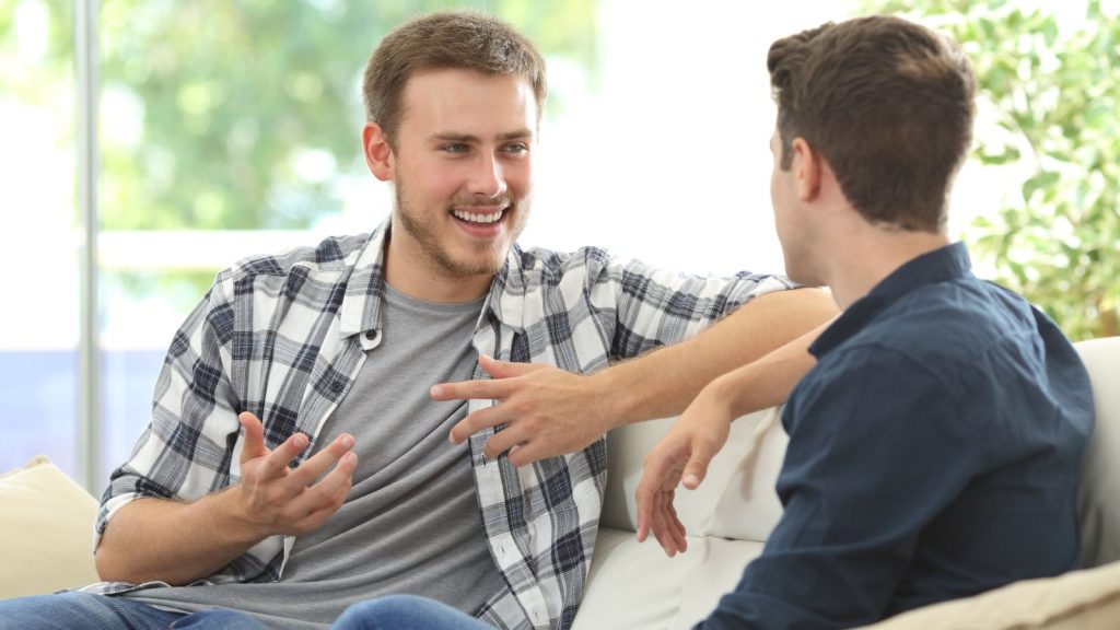 Two friends talking sitting in a couch in the living room with a window in the background at home