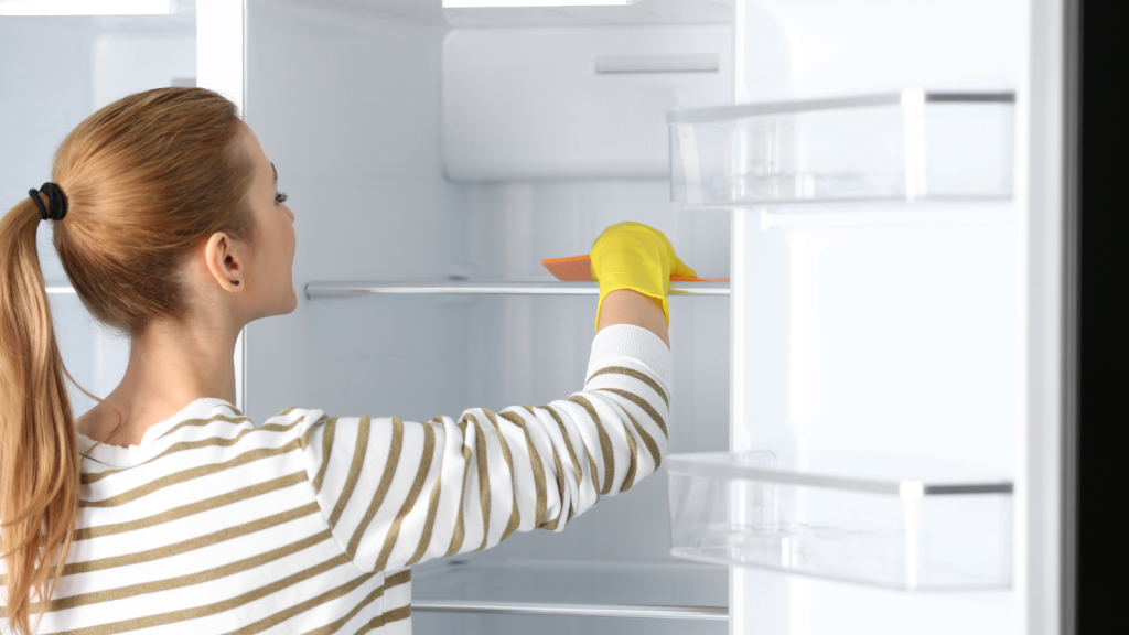 Woman in rubber gloves cleaning empty refrigerator with rag at home