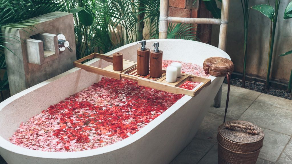 Bathtub with flower petals and beauty products on wooden tray. 