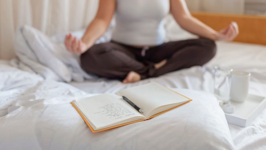 Woman meditating in bed, practicing stress relief challenge with journal about feeling, glass of water, and gratitude list