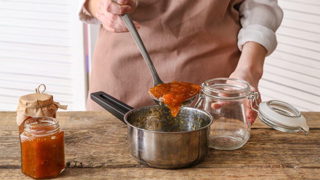 Woman pouring tasty peach jam from saucepan into jar in kitchen.