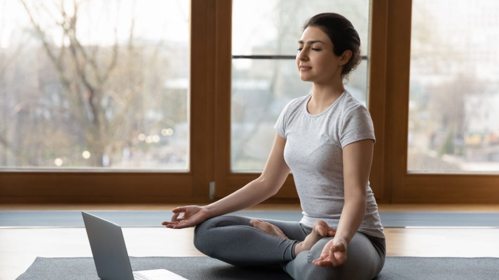 Serene Indian ethnicity woman sit cross-legged on yoga mat listen soothing music on laptop, do meditation practise with eyes closed alone indoor