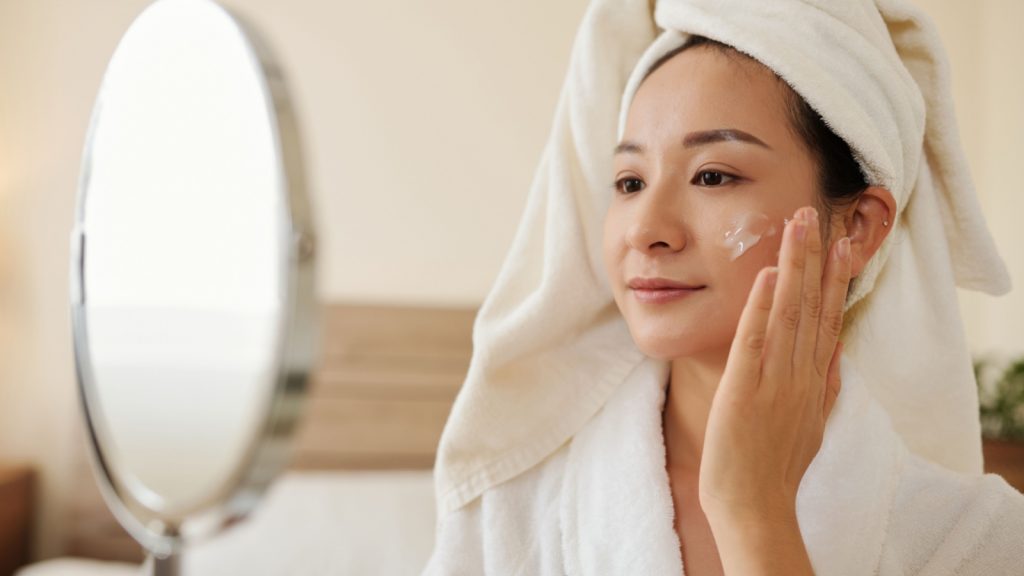 young woman in bathrobe looking at magnifying mirror and applying cream on her face