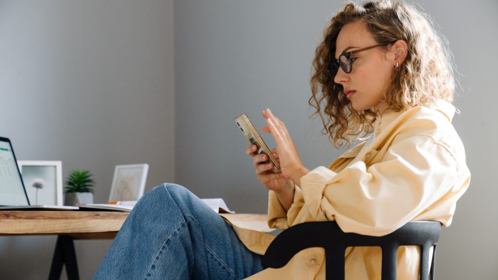 young, curly, serious woman in glasses sitting while scrolling through phone