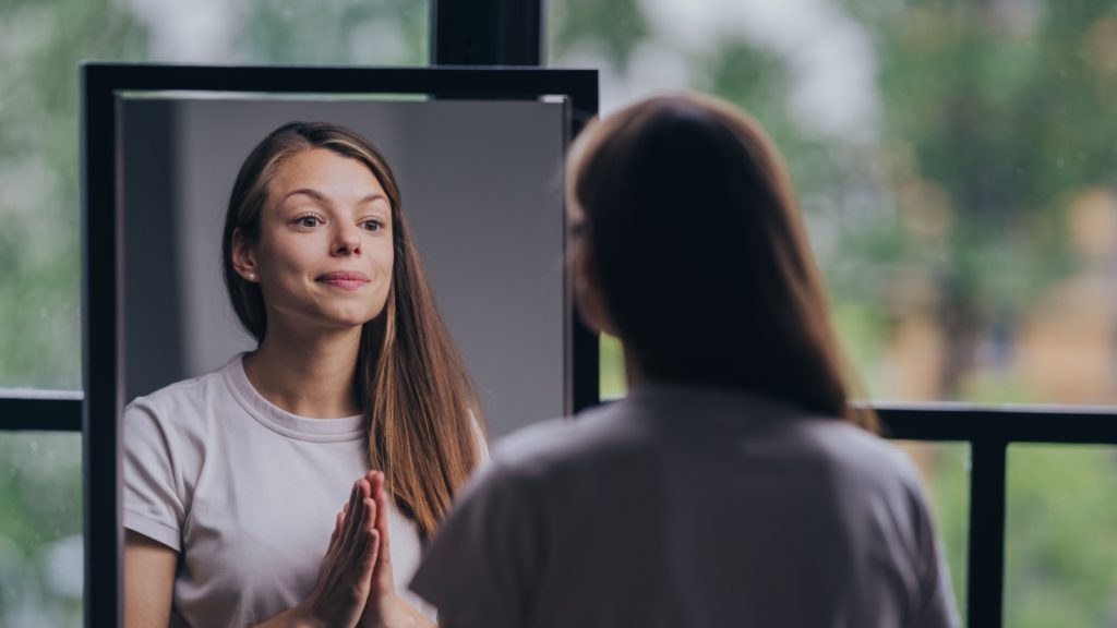 reflective young woman in a casual t-shirt practicing self-affirmation in front of a mirror in a well-lit room