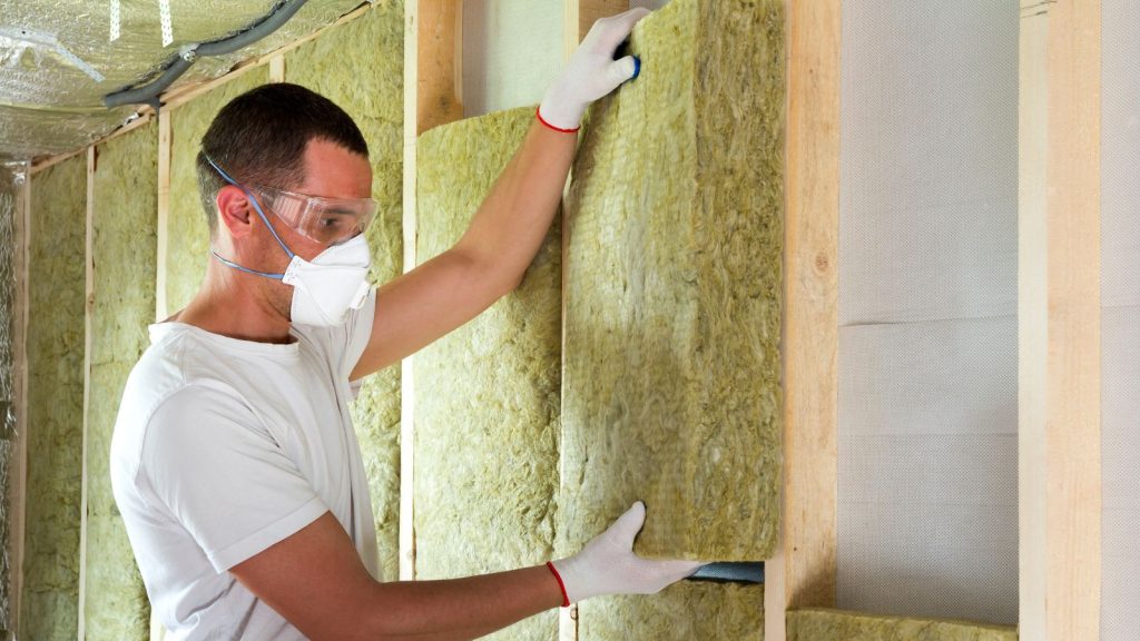 Worker in protective goggles and respirator insulating rock wool insulation in wooden frame.